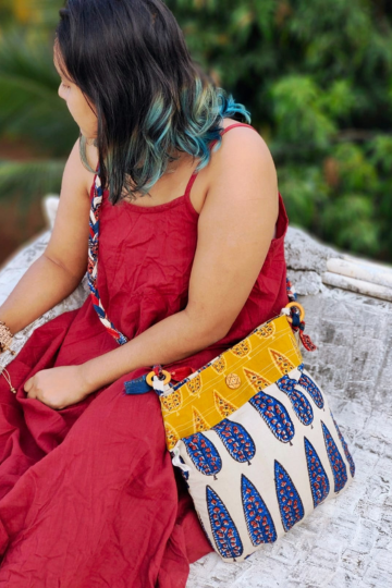 Back view of a woman in a red dress sitting outdoors, showcasing a sustainable, off-white cotton sling bag with blue and orange leaf block prints, and a bold braided strap, crafted by Almitra Sustainables.