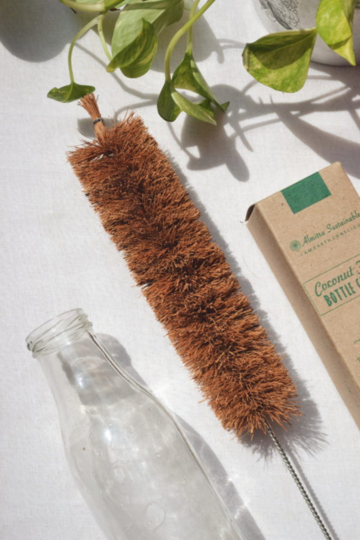 Eco-friendly bottle brush made from coconut coir fibers with a stainless steel handle by Almitra Sustainables, lying beside a reusable glass bottle and recyclable kraft packaging, ideal for sustainable cleaning of bottles and narrow containers.