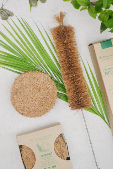 Flat lay of a sustainable kitchen cleaning combo featuring a round coconut coir scrub pad and long bottle brush with coir bristles, placed on palm leaves alongside eco-conscious packaging, representing Almitra Sustainables’ natural zero-waste cleaning products.