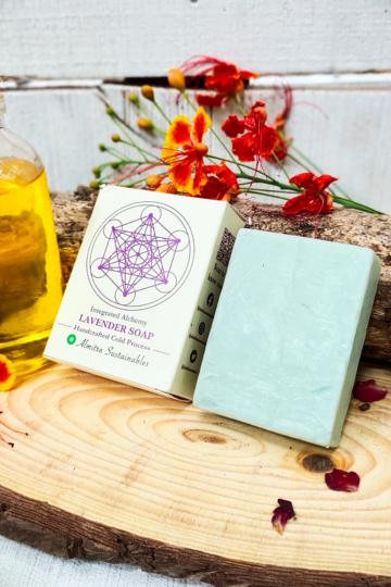Flat-lay of Almitra Sustainables Lavender Aromatherapy Soap with natural wood background, yellow essential oil bottle, and bright tropical flowers – zero-waste packaging, organic ingredients, and soothing fragrance.