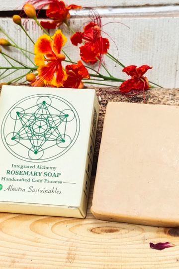 Almitra Sustainables' rosemary cold process soap placed beside eco-friendly packaging adorned with a sacred geometric design, set against a wooden backdrop with vibrant orange flowers.