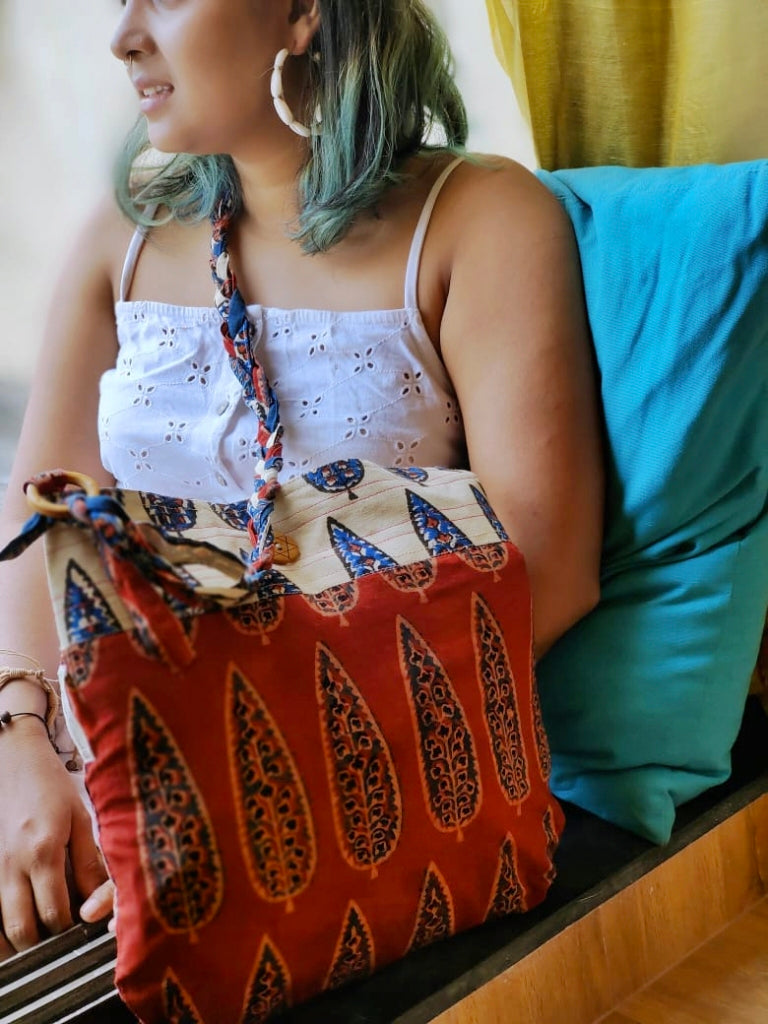 Sustainable fashion portrait of a woman with teal-tinted hair wearing a white eyelet blouse, showcasing a handcrafted maroon and off-white sling bag with earthy Indian-inspired leaf motifs and a braided cotton strap.