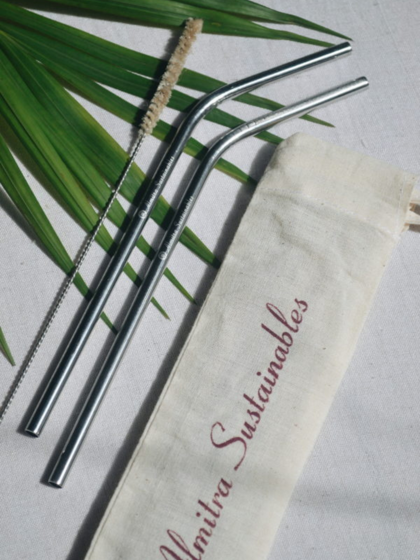 Set of two stainless steel bent straws with a natural bristle cleaning brush and cotton travel pouch printed with Almitra Sustainables branding, laid on a natural palm leaf backdrop – ideal for eco-conscious hydration on the go.