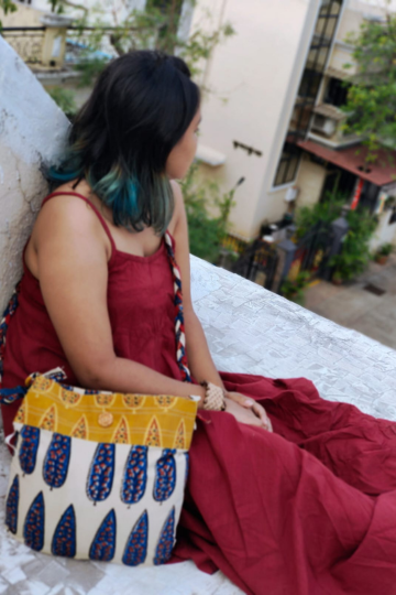 Woman seated on a rooftop wearing a red dress and carrying an Almitra Sustainables block printed sling bag with a braided strap and blue leaf design on off-white cotton.