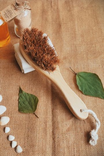Top-down shot of Almitra Sustainables’ coconut coir dry brush, placed with handmade bath salts and oil bottles on a jute background, showcasing its natural and plastic-free design.