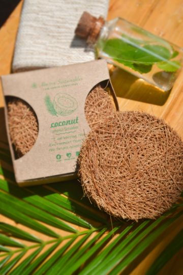 Top-down image showing the round coconut fiber scrubber and open box pack of 3 alongside natural elements like a palm leaf, handmade soap, and herbal oil – highlighting eco-conscious cleaning essentials.