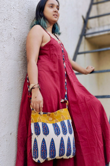 Model in a flowy red dress standing outdoors with a sustainable sling bag featuring a braided strap and blue-and-mustard ethnic print, designed by Almitra Sustainables for conscious everyday fashion.