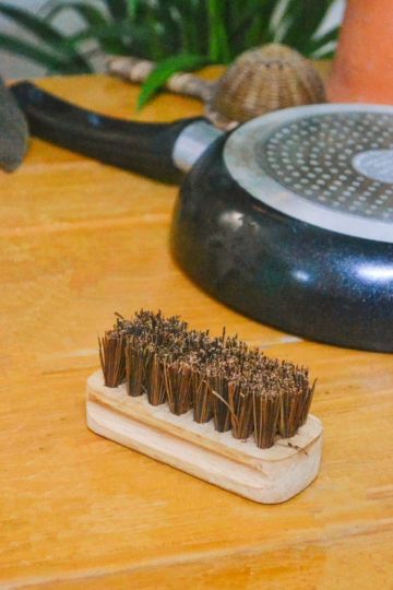 Flat lay of the eco-conscious kitchen scrub brush by Almitra Sustainables, lying on a wooden table next to a stainless steel pan base, emphasizing its compact, ergonomic design.