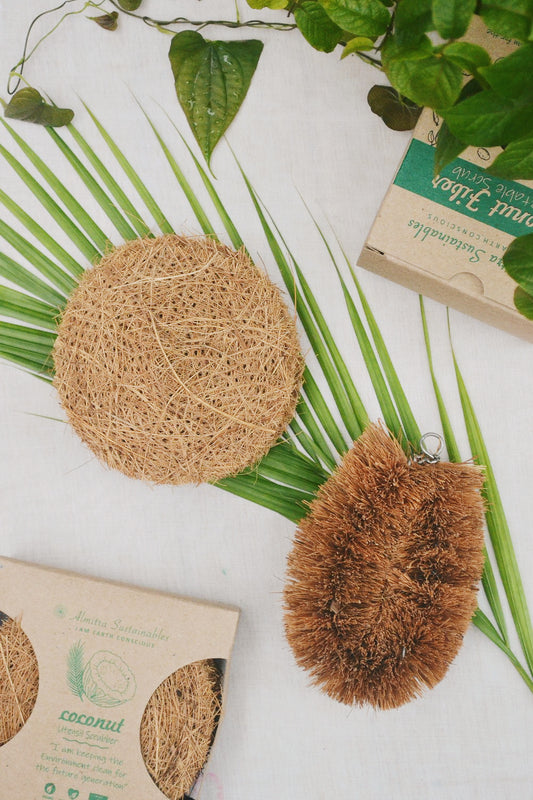 A flat lay of an eco-friendly kitchen cleaning set featuring a round coconut coir scrubber and a vegetable brush made from natural coconut fibers, placed on palm leaves with green foliage and sustainable packaging, reflecting Almitra Sustainables’ commitment to zero-waste living.
