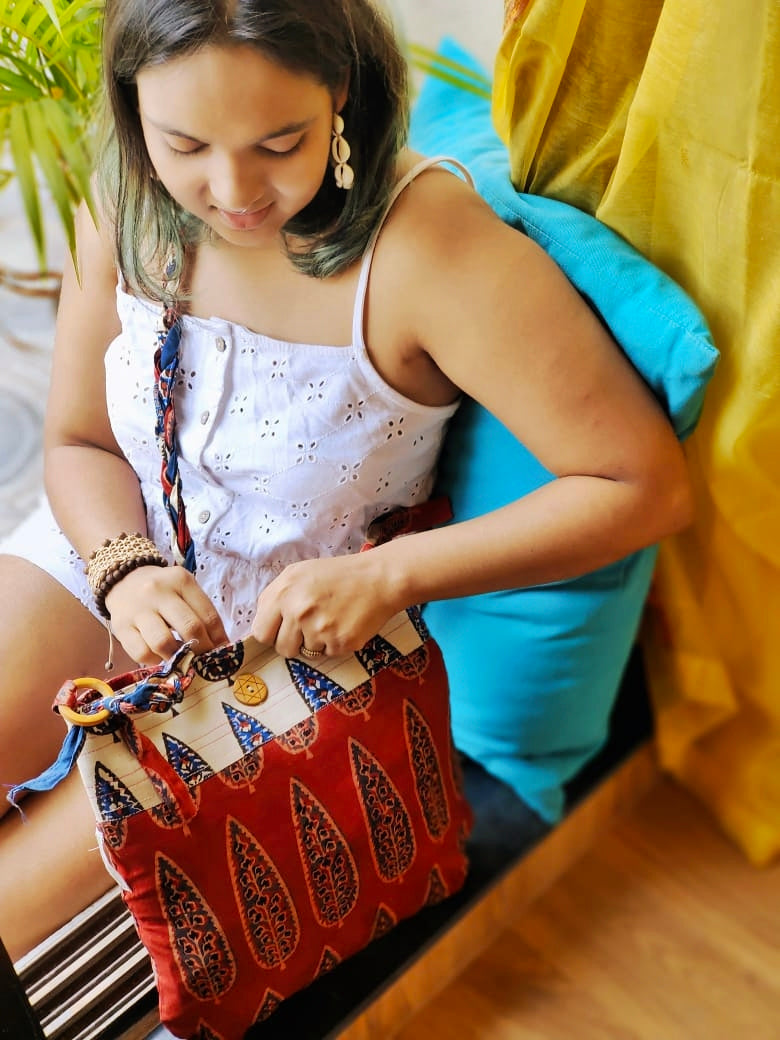 Close-up of a woman in a sleeveless white top opening a maroon and off-white ethnic cotton sling bag with eco-friendly braided strap and natural block print design from Almitra Sustainables.