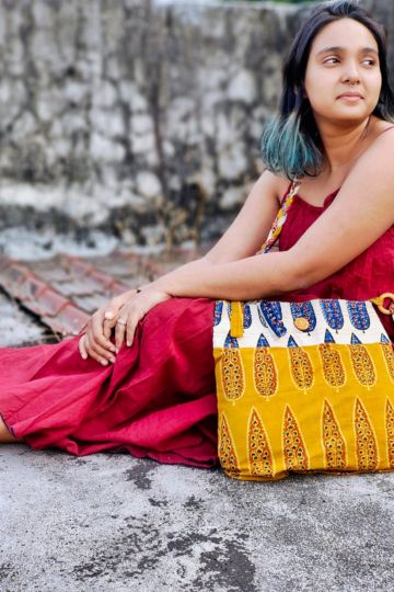 Woman in a maroon cotton dress sitting outdoors with a yellow and off-white hand block printed sling bag from Almitra Sustainables, featuring braided straps and traditional Indian motifs.