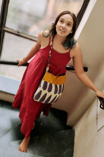 Young woman in a red dress standing on stairs, wearing a handmade ethnic sling bag with blue block-printed leaf motifs and a mustard yellow border, promoting Almitra Sustainables’ eco-conscious, artisan-crafted accessories.