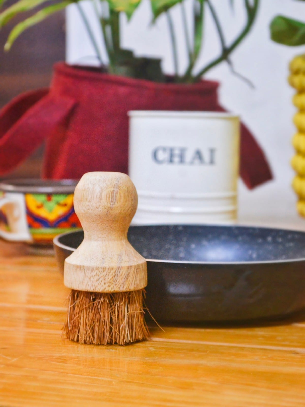 A handcrafted wooden dish scrubber with dense coconut coir bristles standing upright on a wooden kitchen counter. Behind it sits a non-stick pan and cozy kitchen decor elements like a "CHAI" container and colorful mugs, promoting a zero-waste, sustainable lifestyle.