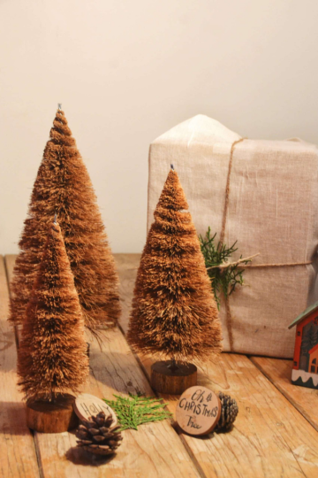 Three eco-friendly coir fiber Christmas trees arranged with a pine cone, green sprig, and a linen-wrapped present, styled as a plastic-free holiday décor setting by Almitra Sustainables.