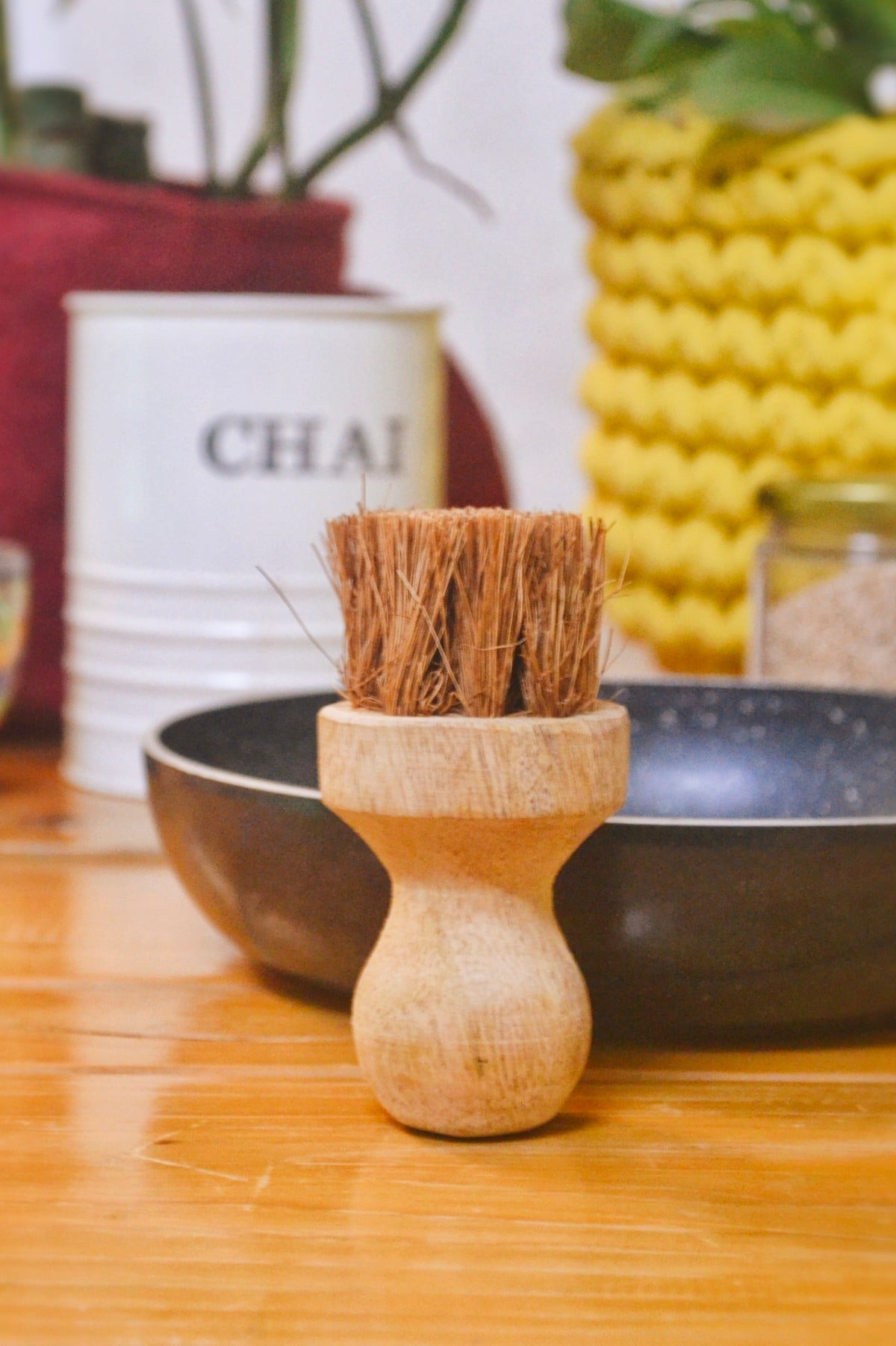 A close-up of an eco-friendly coconut coir dish scrubber with a smooth, ergonomic wooden handle, placed inside a black non-stick frying pan. The background features rustic home decor including a "CHAI" tin and hand-knitted plant holder, evoking a warm, sustainable kitchen vibe.