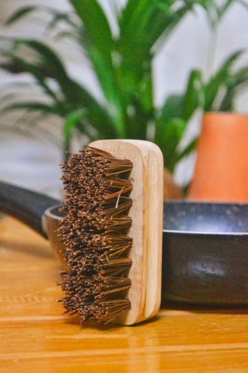 Eco-friendly hard scrub brush with natural coir bristles and a wooden handle, standing vertically on a kitchen counter near a black iron pan, with green indoor plants in the background.