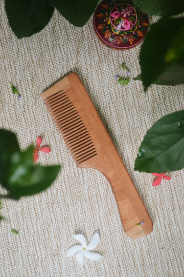 Eco-friendly neem wood comb with handle displayed on a woven mat surrounded by green leaves and tropical flowers, showcasing its smooth texture and zero-waste appeal.