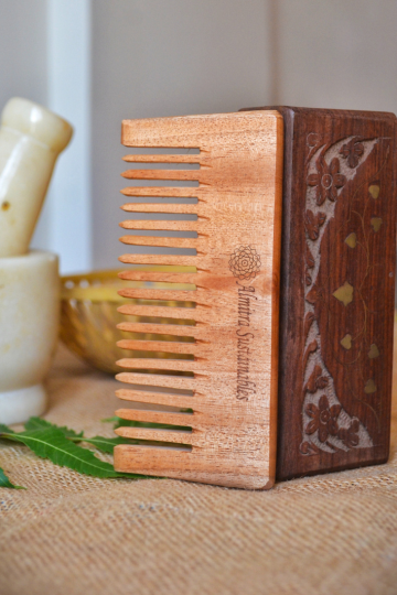 Eco-friendly wide-tooth neem wood comb resting against an intricately carved wooden box, paired with neem leaves and a marble mortar, reflecting its Ayurvedic and scalp-friendly properties from Almitra Sustainables.