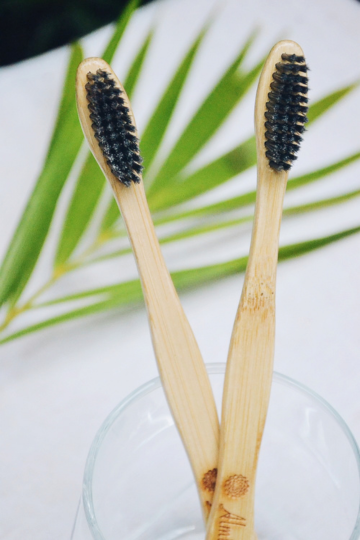 Two bamboo charcoal toothbrushes in a glass jar with green leaf background, minimalist zero-waste bathroom aesthetic