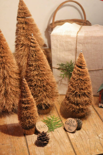 A cluster of handcrafted coir Christmas trees in varying sizes with rustic wooden bases, pinecone, "Holly" and "Oh Christmas Tree" wooden tags, set beside a sustainable cloth-wrapped gift box.