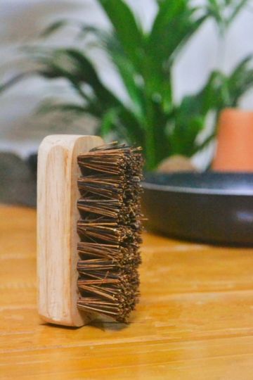 Handcrafted wooden kitchen scrub brush featuring coarse natural bristles, positioned vertically on a wooden table with cookware and eco-inspired decor in the backdrop.