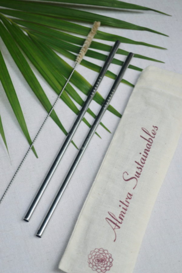 Flat lay of a reusable stainless steel straw set by Almitra Sustainables, featuring two eco-friendly metal straws, a straw cleaning brush, and a washable organic cotton pouch, placed over a green palm leaf for a natural, sustainable aesthetic.
