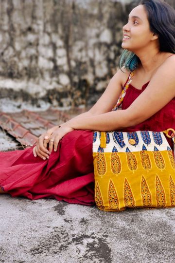 Young woman in a red dress with an Almitra Sustainables hand block printed sling bag in mustard yellow and cream, styled with bold Indian-inspired patterns and an artisan-crafted braided strap.
