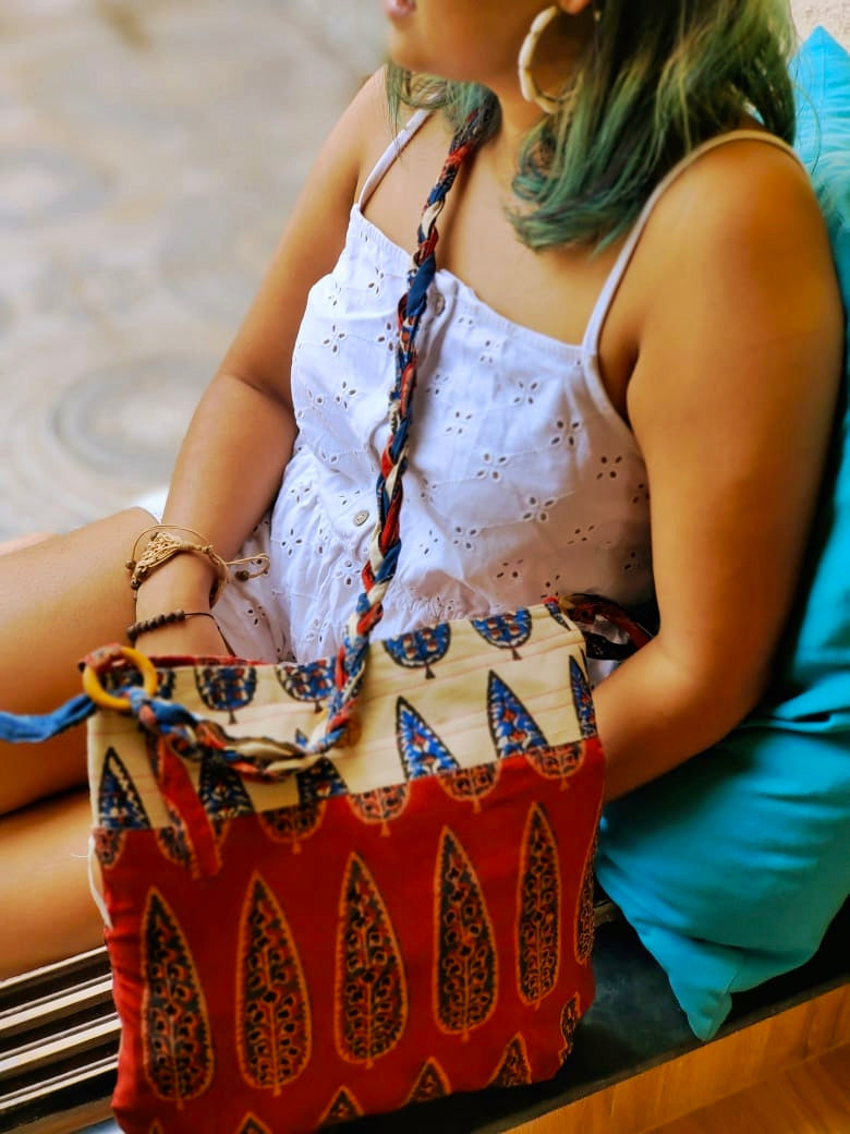 Young woman resting by a window with a maroon and off-white cotton sling bag adorned in traditional block prints and a multicolored hand-braided shoulder strap, part of Almitra Sustainables' eco-conscious collection.