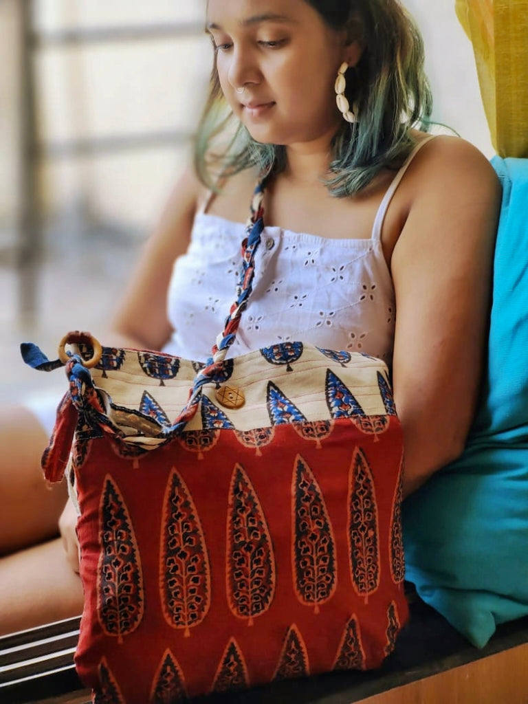 Woman seated indoors wearing a white eyelet cotton top, holding a maroon and off-white hand block printed cotton sling bag by Almitra Sustainables, featuring traditional Indian motifs and a braided strap.