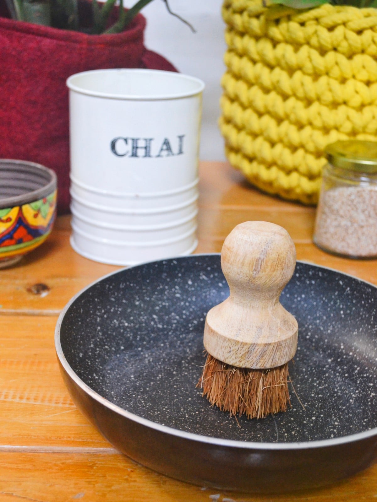 A plastic-free, coconut bristle kitchen scrubber featuring a curved wooden handle, standing on a wooden counter with a frying pan and knitted planter in the background. Designed for eco-conscious households embracing sustainable and vegan kitchen practices.
