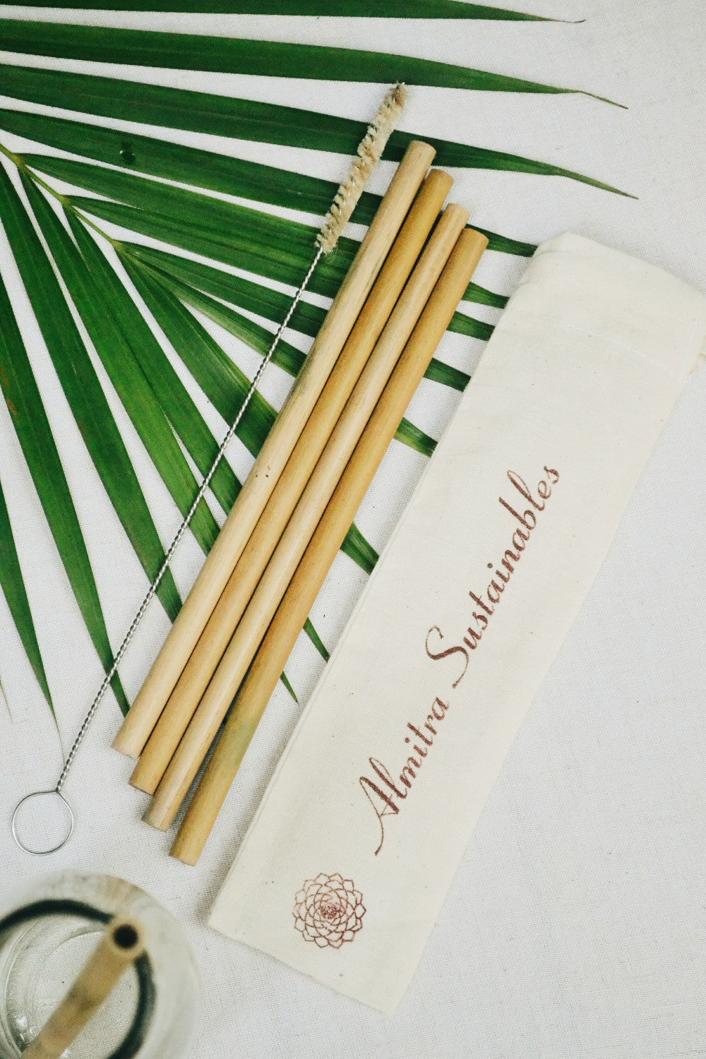 A set of reusable bamboo straws alongside a cleaning brush and a cloth bag with the brand name 'almitra sustainables' printed on it, placed on a white surface with green palm leaves in the background.