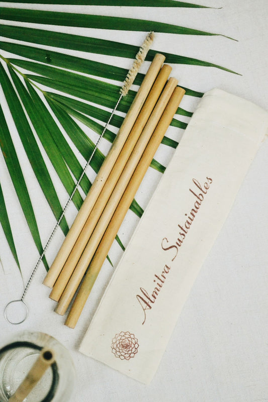 A set of reusable bamboo straws alongside a cleaning brush and a cloth bag with the brand name 'almitra sustainables' printed on it, placed on a white surface with green palm leaves in the background.