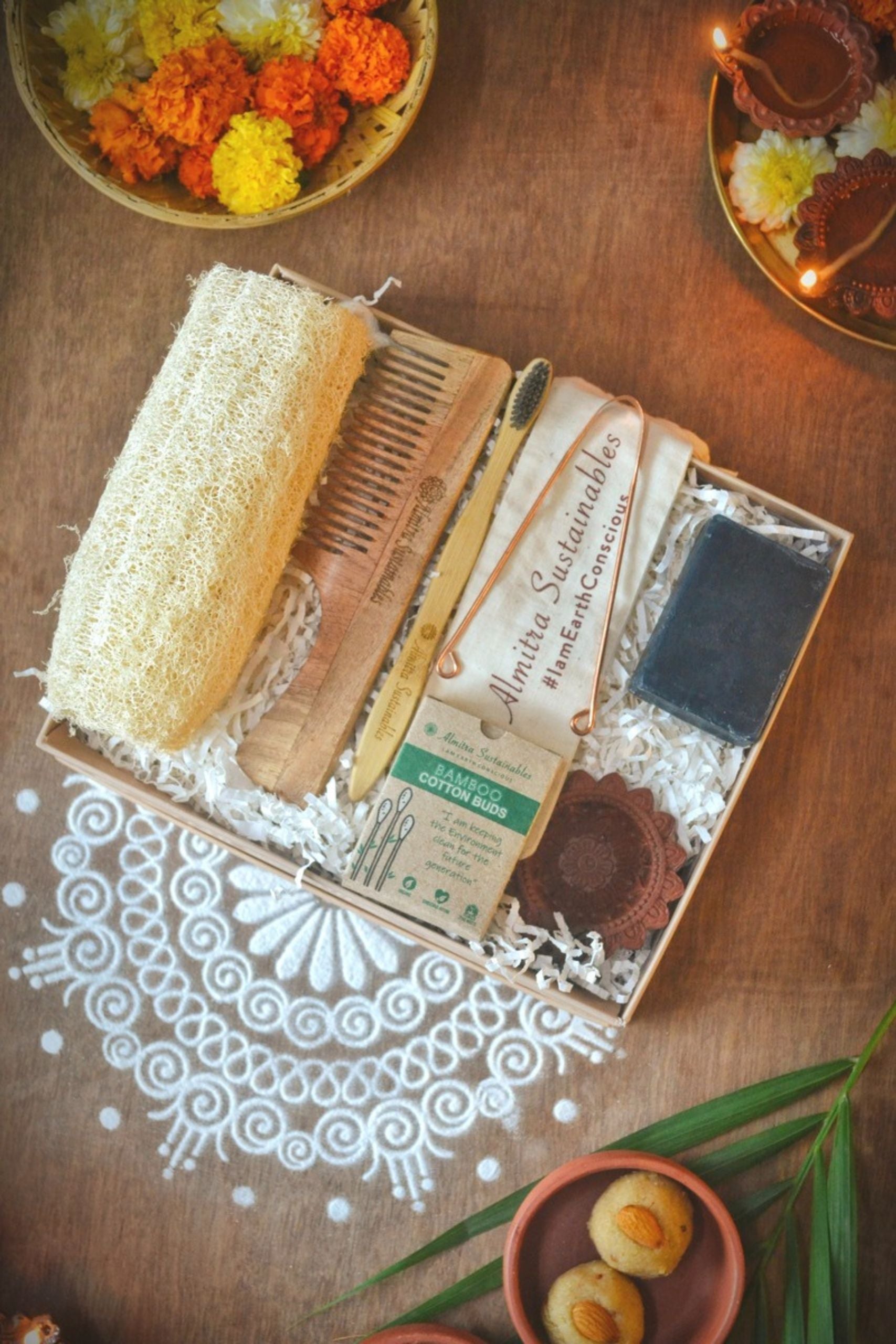 Eco-friendly self-care gift hamper by Almitra Sustainables containing a natural loofah, bamboo toothbrush, wooden comb, handmade soap, bamboo cotton buds, and copper tongue cleaner, placed on a festive wooden background with flowers and diyas — sustainable gifting box for Diwali or wellness occasions.