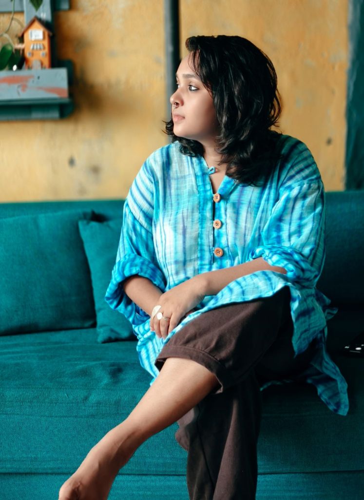Woman sitting on a teal couch wearing a blue tie-dye shirt and brown pants.