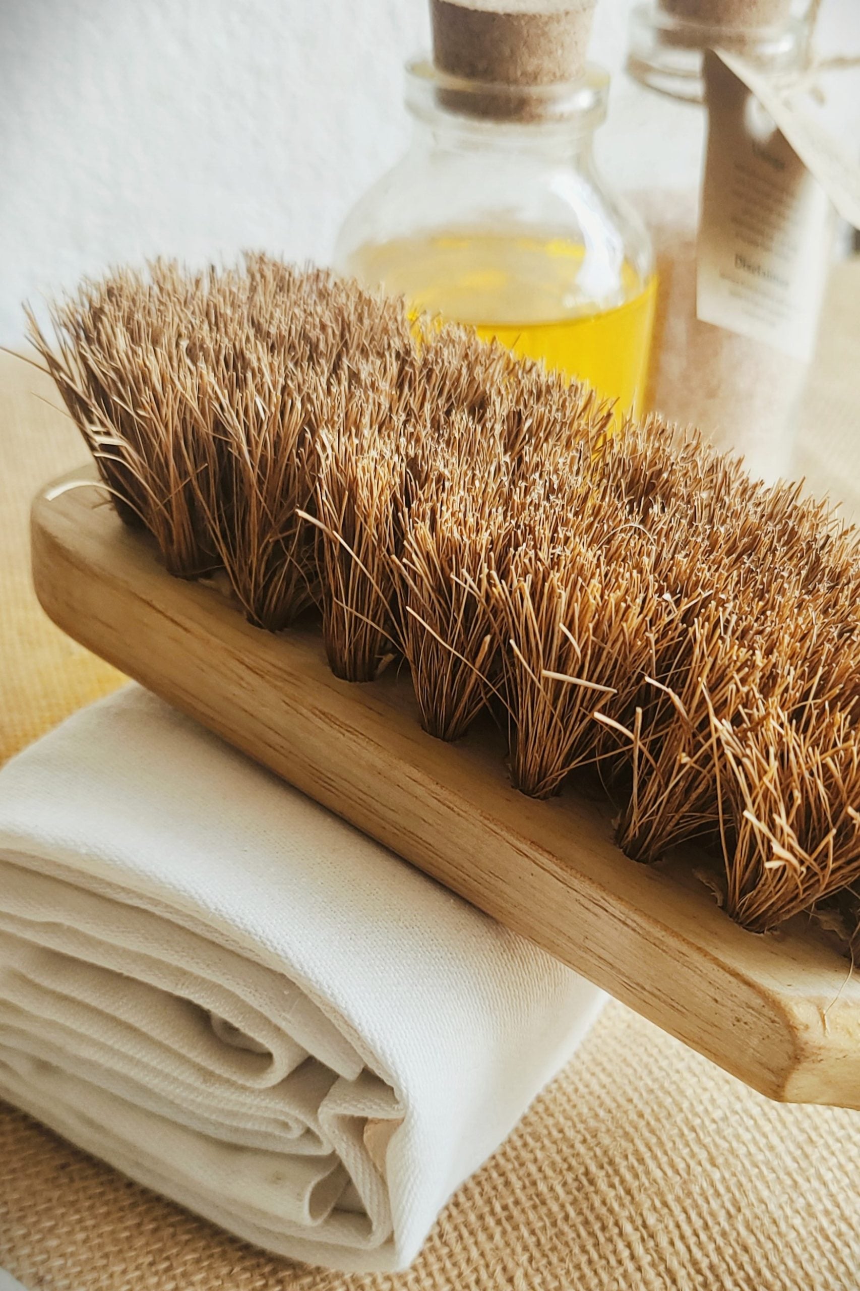 Close-up of a natural coconut coir body brush with a bamboo wood handle, resting on a rolled organic cotton towel beside glass bottles of essential oils — perfect for eco-conscious exfoliation and dry brushing.

