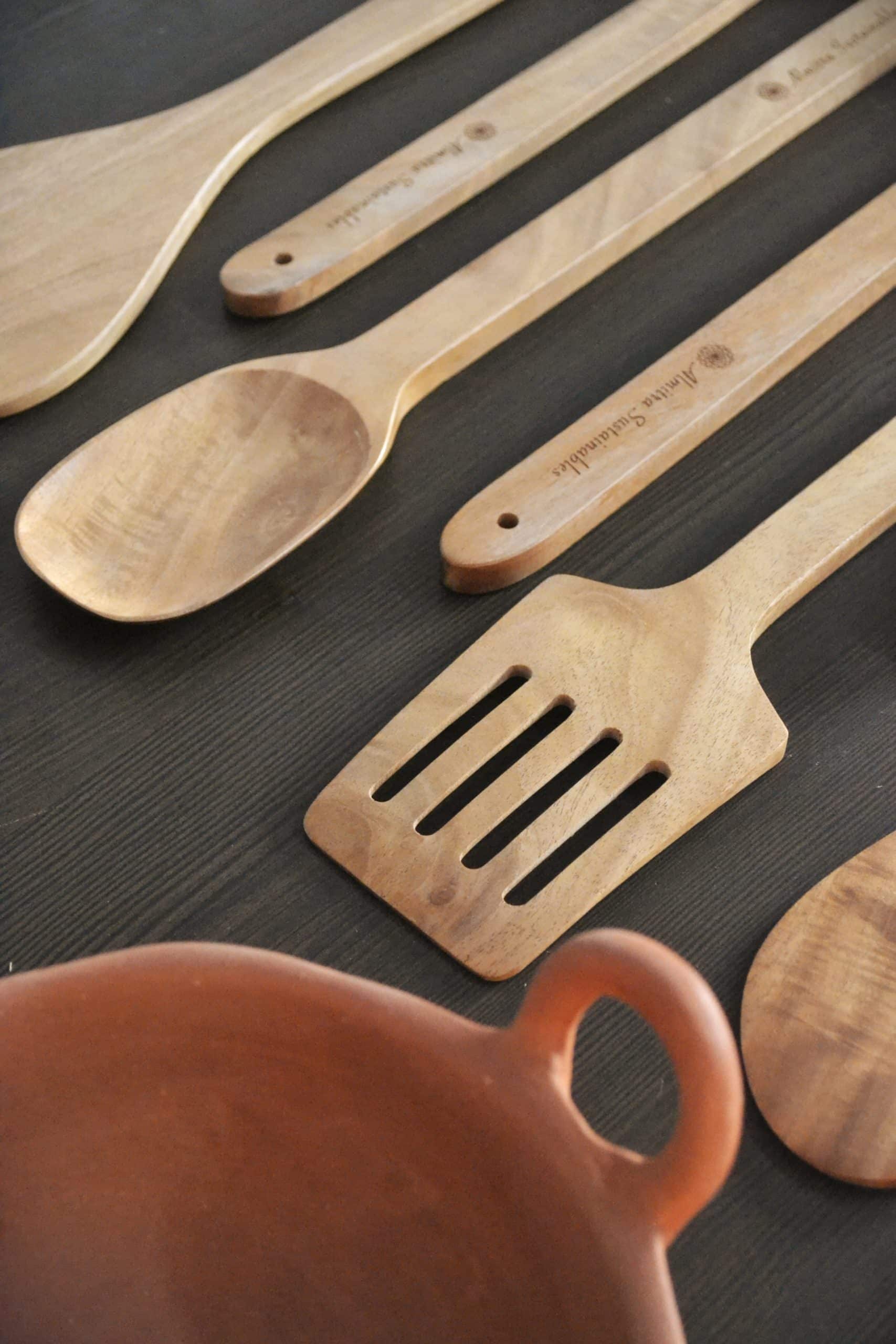 Close-up of sustainable neem wood kitchen utensils from Almitra Sustainables, showcasing fine wood grain textures and engraved handles with the brand name, laid out on a dark countertop with a terracotta bowl partially in view.

