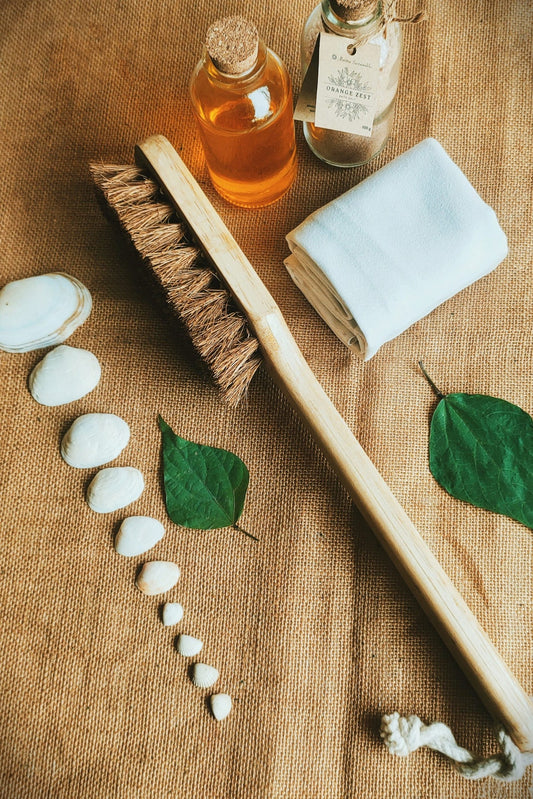 Flatlay of Almitra Sustainables' eco-friendly bath essentials including a long-handled coconut coir body brush, rolled cotton towel, glass bottles of orange zest oil and bath salt, green leaves, and white seashells — a complete sustainable spa gift set.

