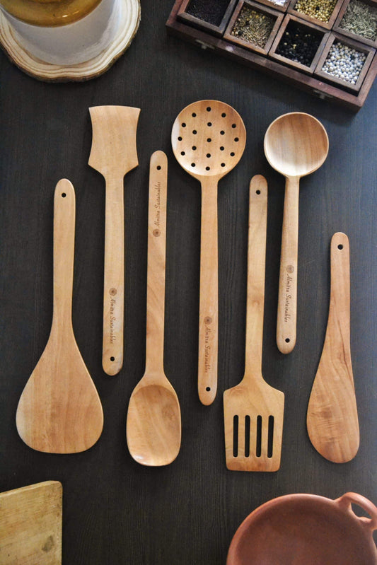 "Eco-friendly handcrafted wooden kitchen utensils set by Almitra Sustainables, including spatula, ladle, slotted spoon, and turner made from sustainable wood, displayed on a dark countertop."