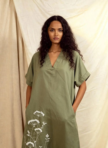 Woman wearing a green dress with white floral embroidery against a beige curtain background