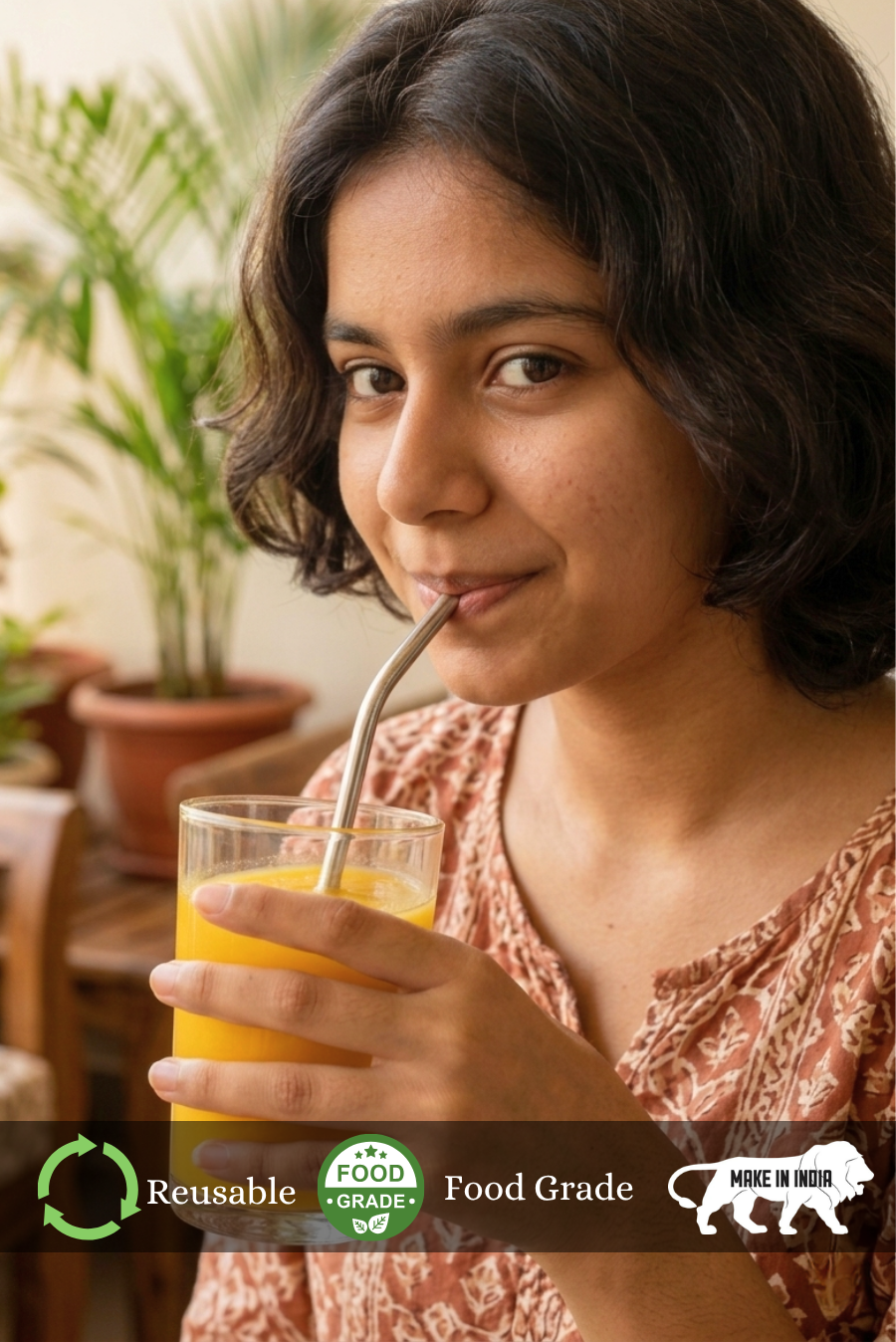 Woman drinking orange juice from a glass with a reusable straw of Almitra Suatainables, set against a blurred indoor background.