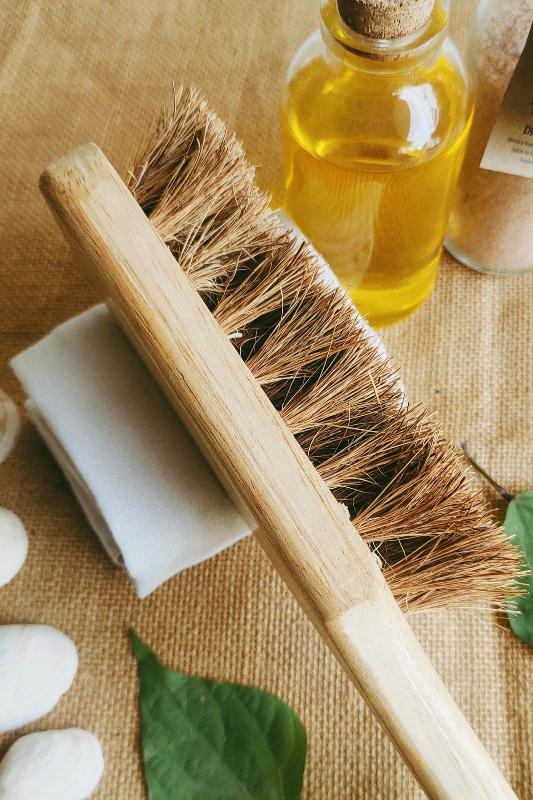 Handmade bath brush with coconut fiber bristles and a bamboo handle, styled with glass jar of essential oil, cotton towel, and green leaves — sustainable and biodegradable skincare accessory.


